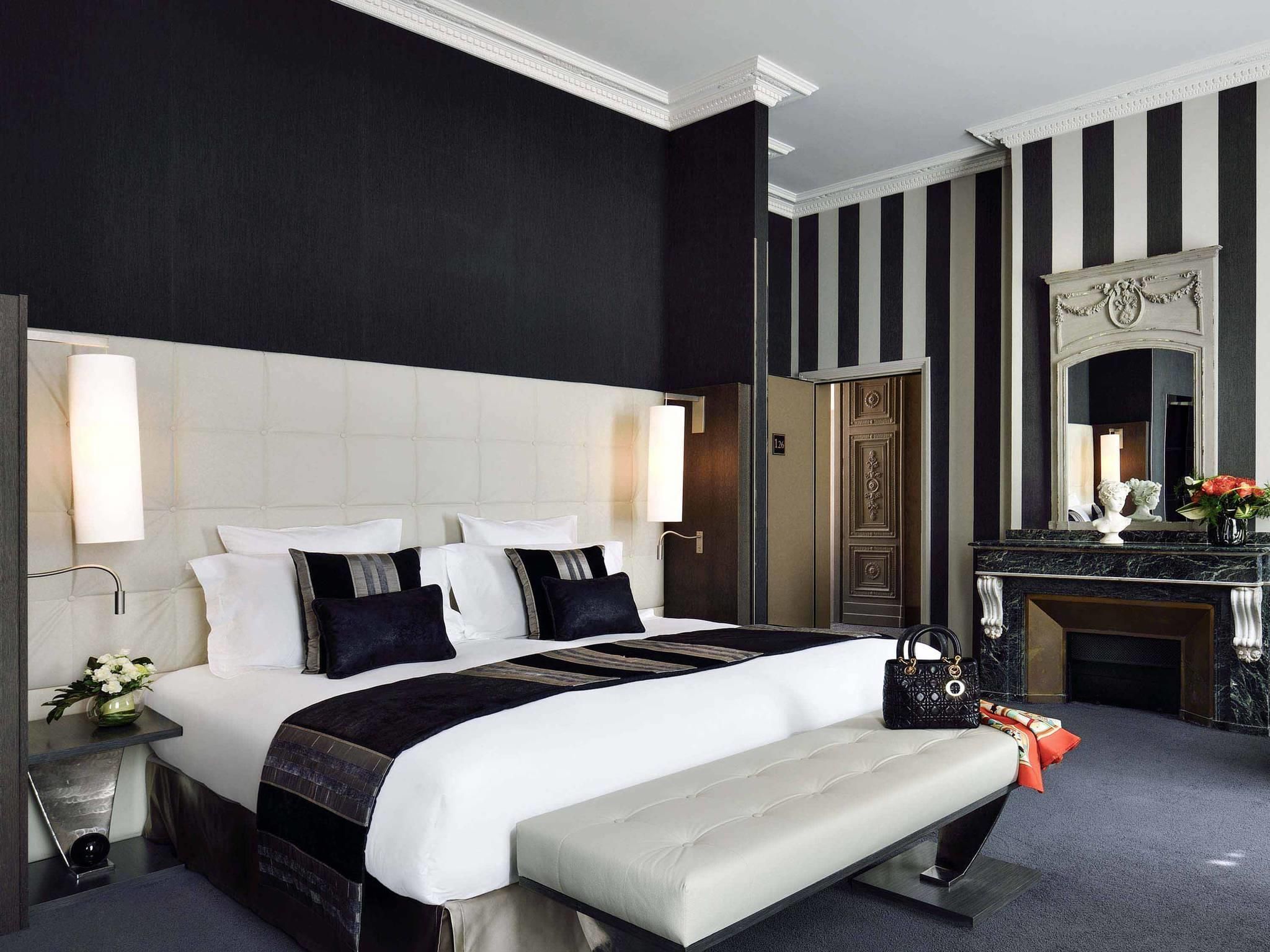 Quarto de hotel com uma cama grande, uma mesa de cabeceira com luminária, um banco, cômoda e um espelho.