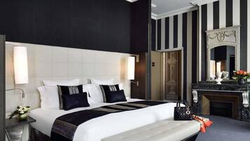 Quarto de hotel com uma cama grande, uma mesa de cabeceira com luminária, um banco, cômoda e um espelho.