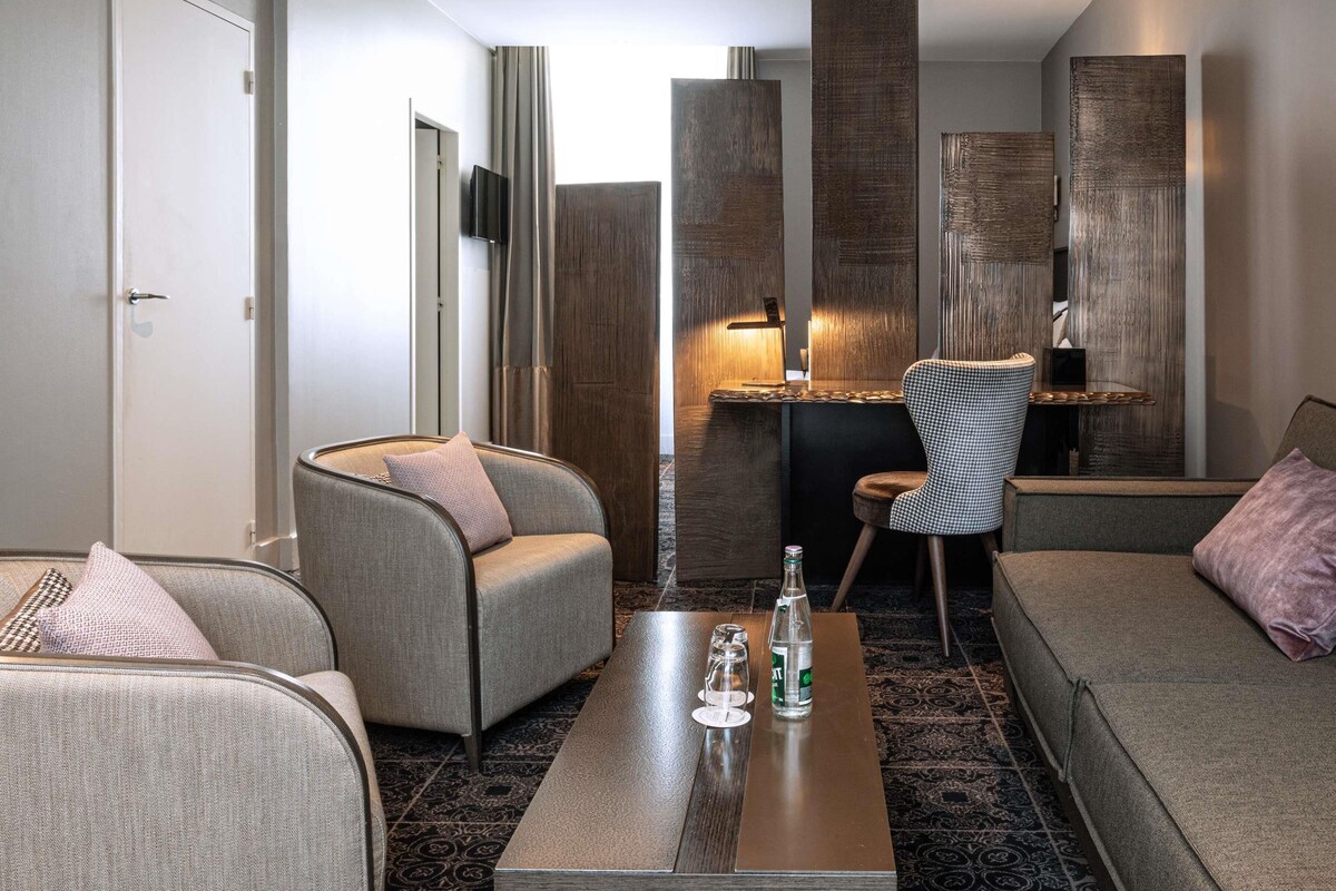A modern hotel room with a sofa, armchair, and a small table with a bottle and glasses.