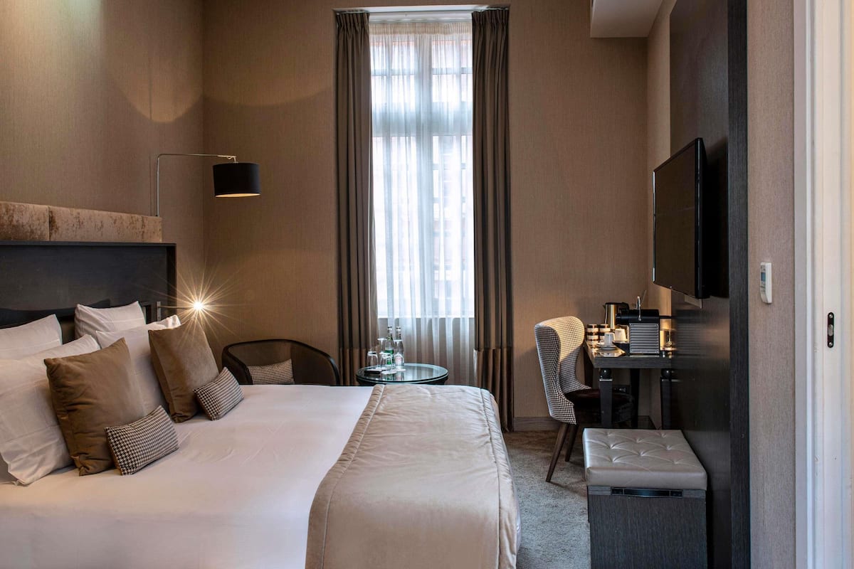 Ein Hotelzimmer mit einem großen Bett, einem Schreibtisch mit Stuhl, einem Fernseher und einem Fenster mit Vorhängen.