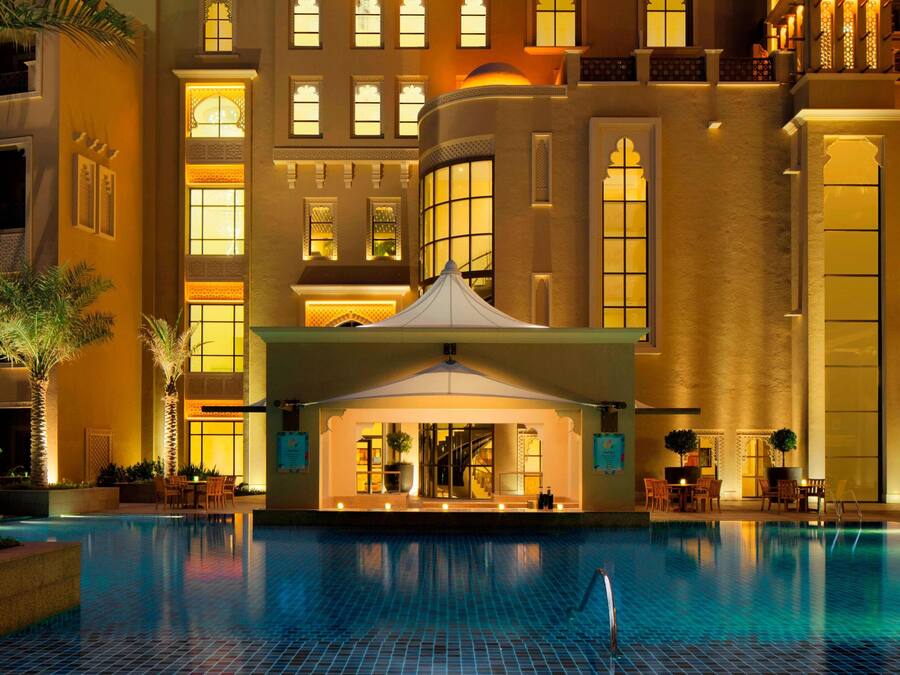 Un espace piscine d’hôtel doté d’un coin salon abrité, de palmiers et d’un grand bâtiment avec de nombreuses fenêtres et des balcons.