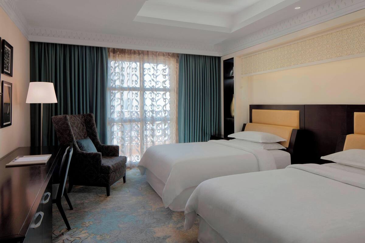 Une chambre d’hôtel avec deux lits, un bureau, une chaise et une grande fenêtre avec des rideaux.