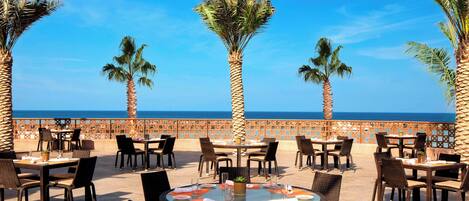A seaside restaurant with outdoor seating, palm trees, and a view of the ocean.