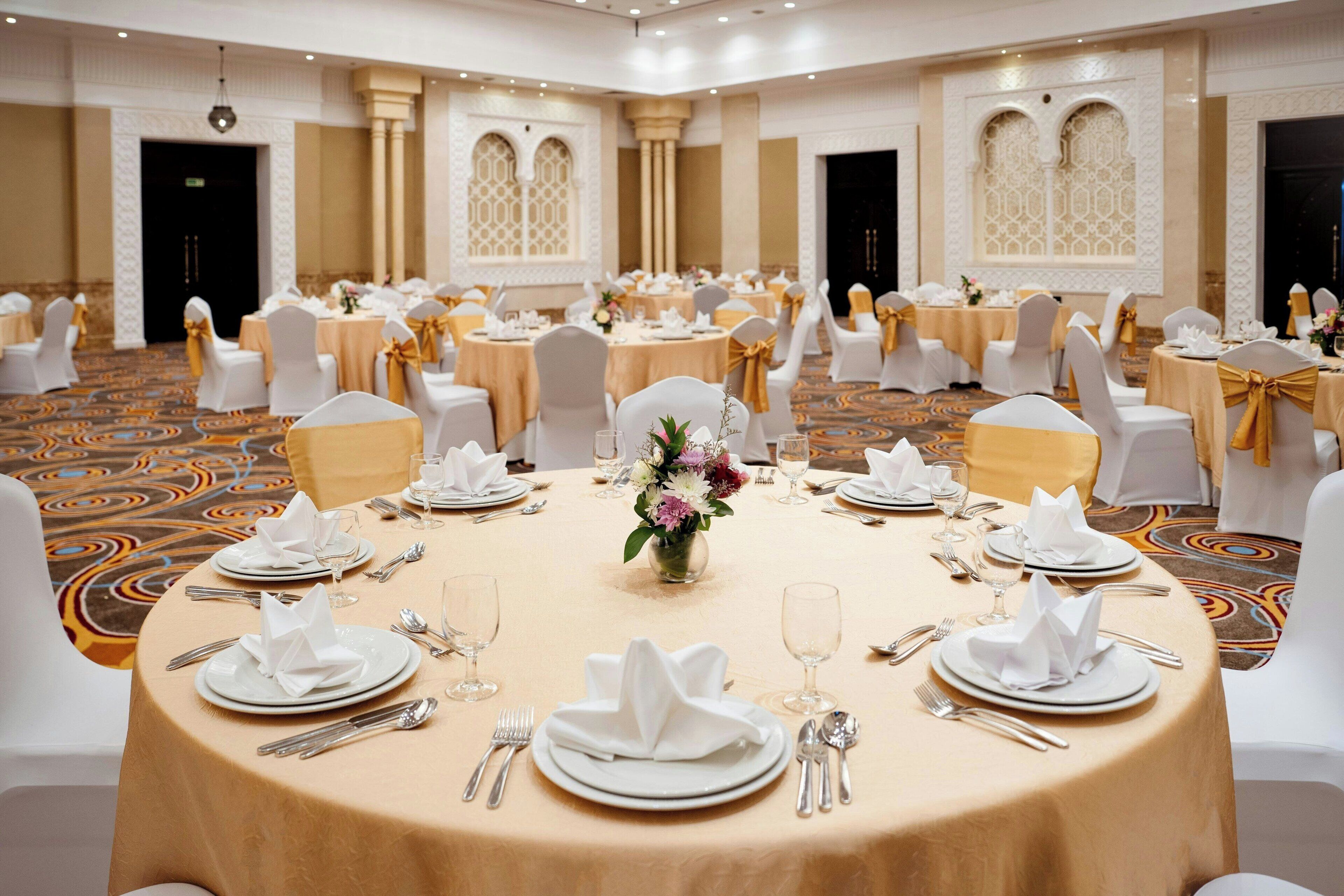A banquet hall with round tables set for a formal event, featuring white tablecloths, gold sashes, and floral centerpieces.