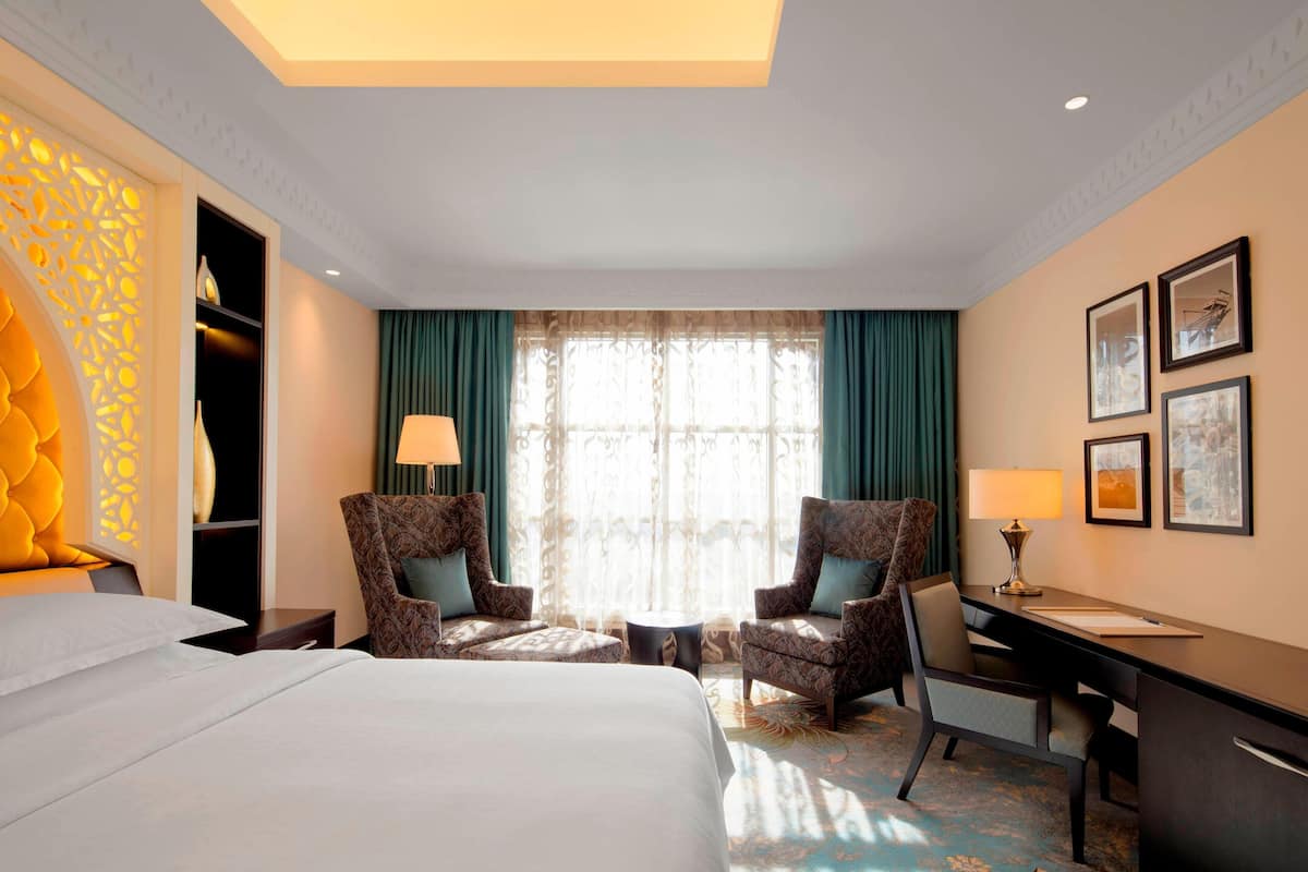 Une chambre d’hôtel avec un grand lit, deux fauteuils, un bureau et une fenêtre avec des rideaux.