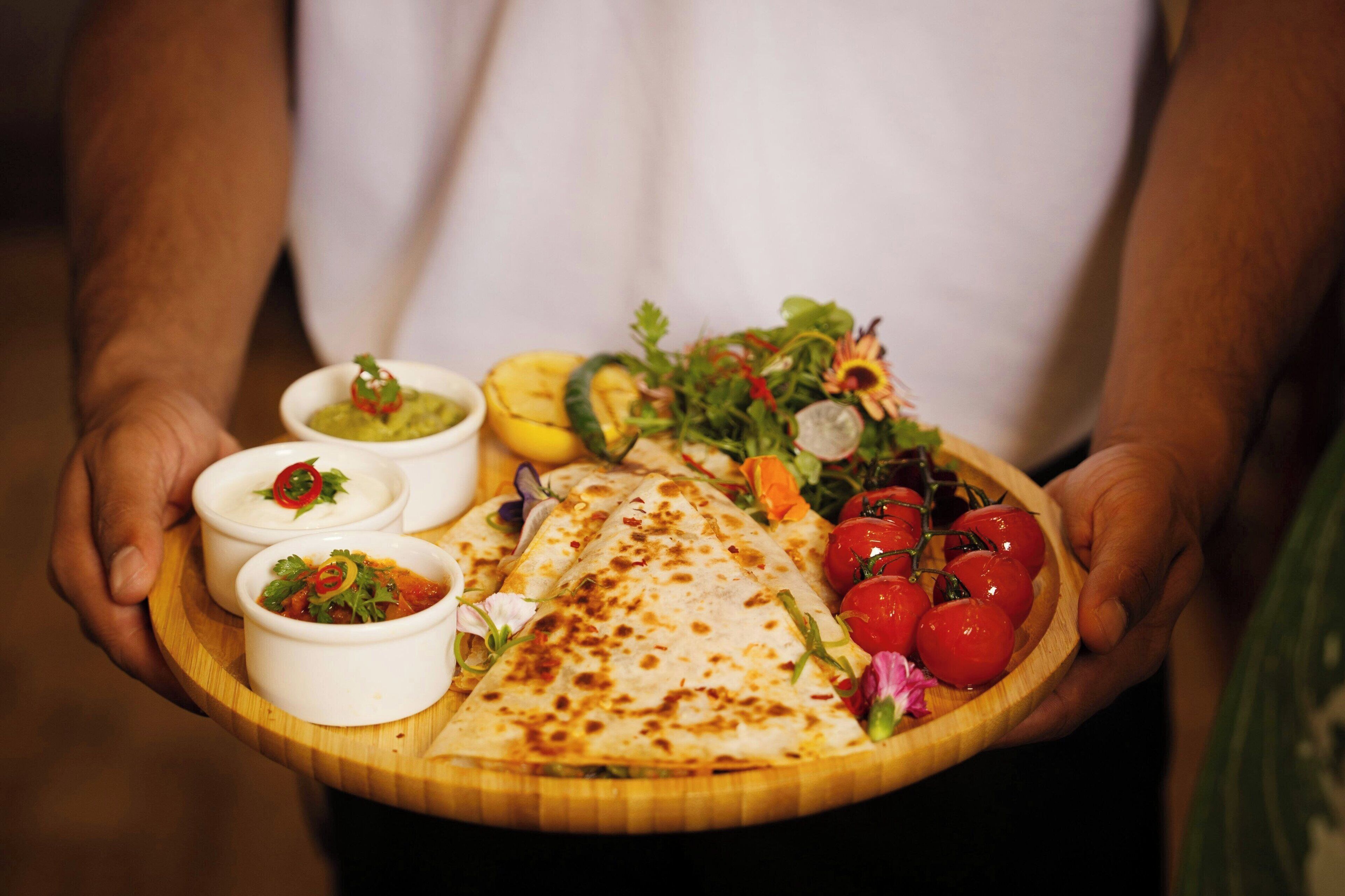 A person holding a wooden board with a quesadilla, salad, and three small bowls of dips.