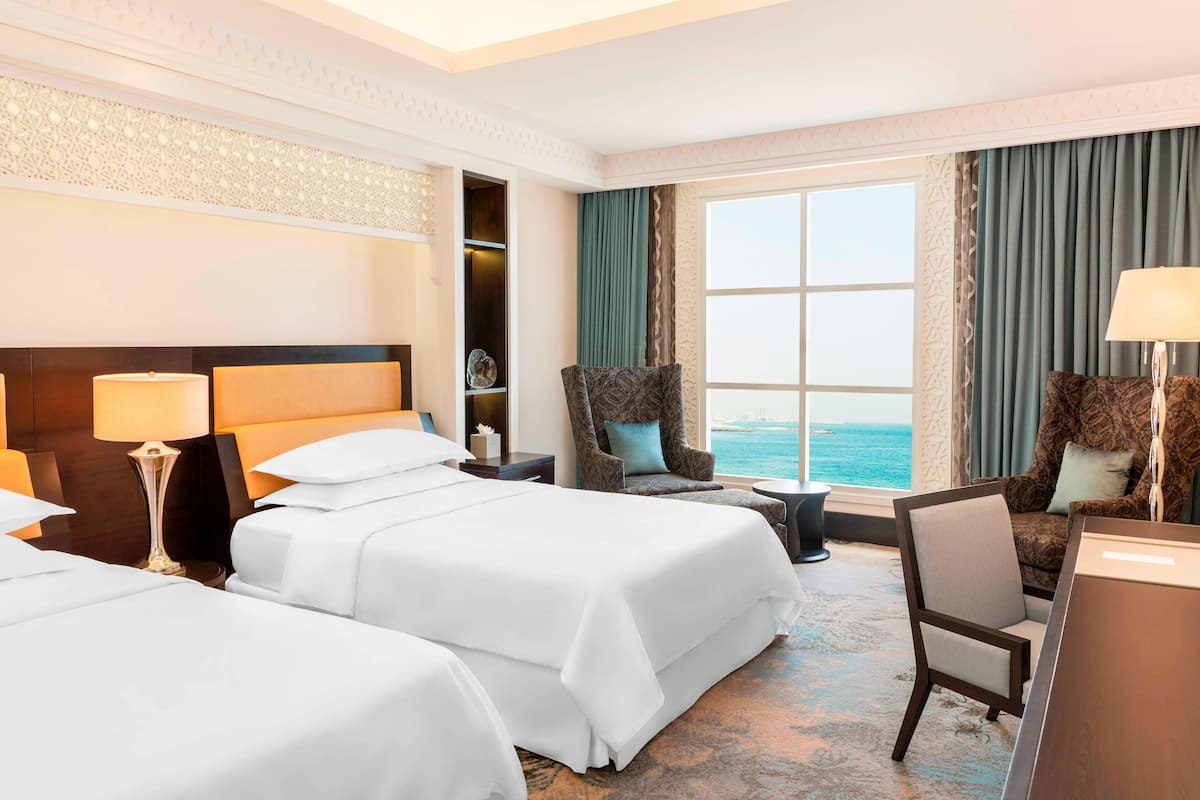 Une chambre d’hôtel avec deux lits, un coin salon donnant sur la mer et un bureau avec une lampe.