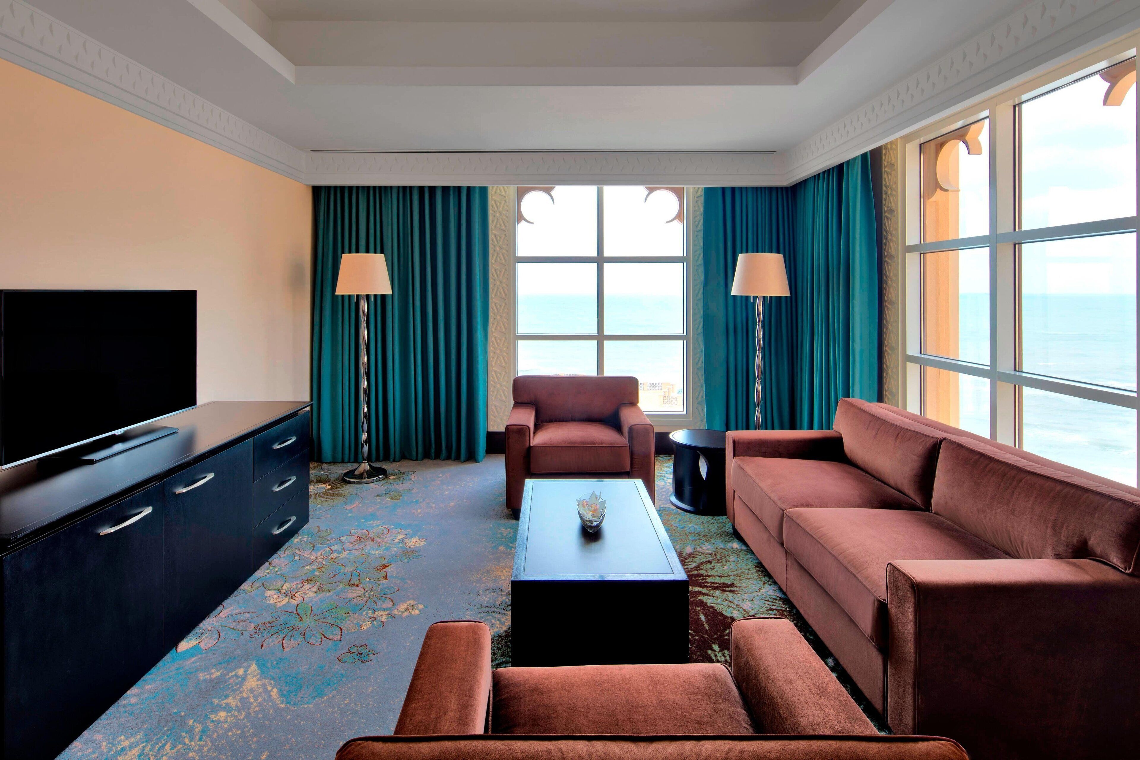 A modern hotel room with a large flat-screen TV, a sofa, a loveseat, and a coffee table. There are two lamps and a view of the ocean through large windows.