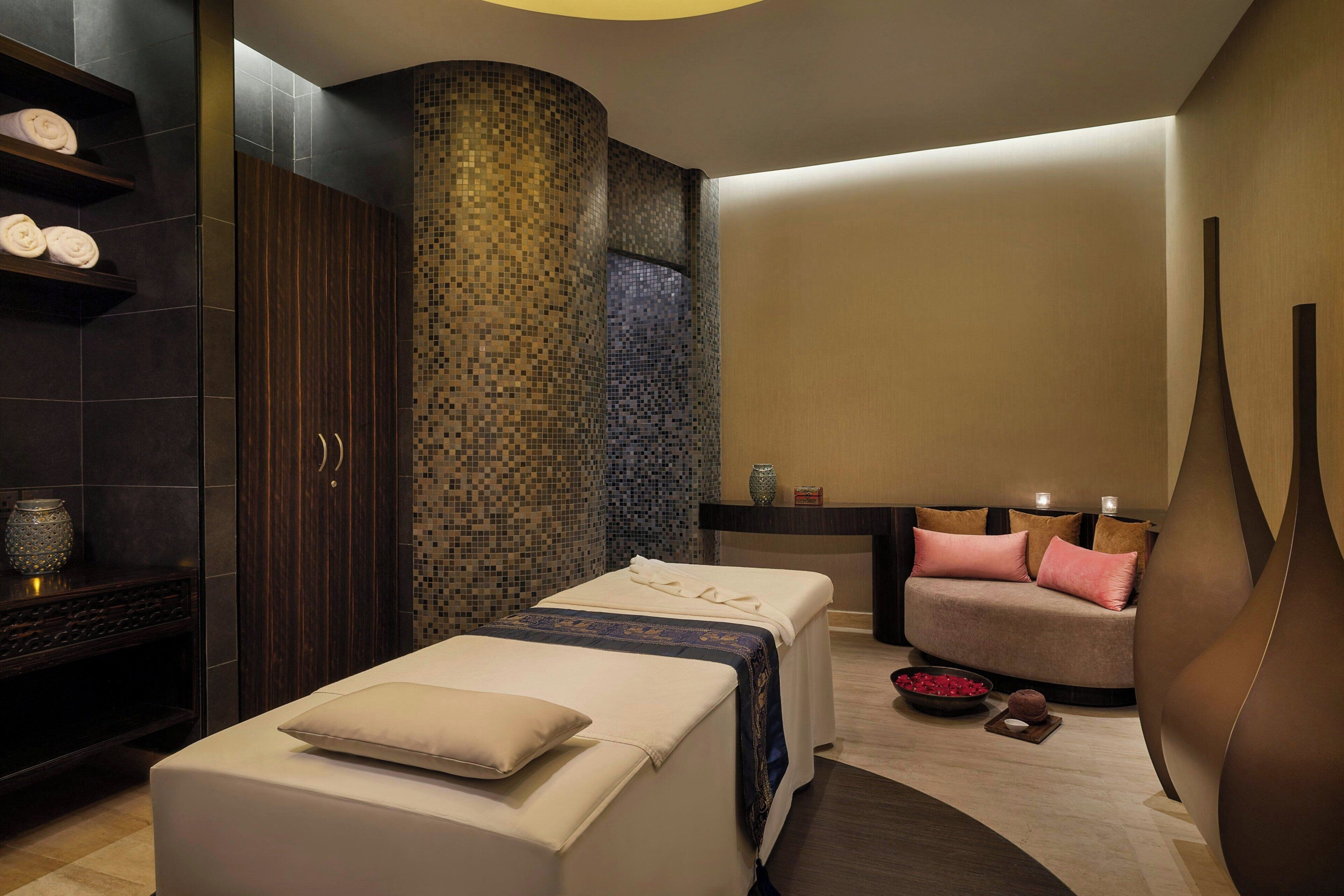 A spa room with two massage tables, a seating area with cushions, a decorative vase, and a tiled wall.