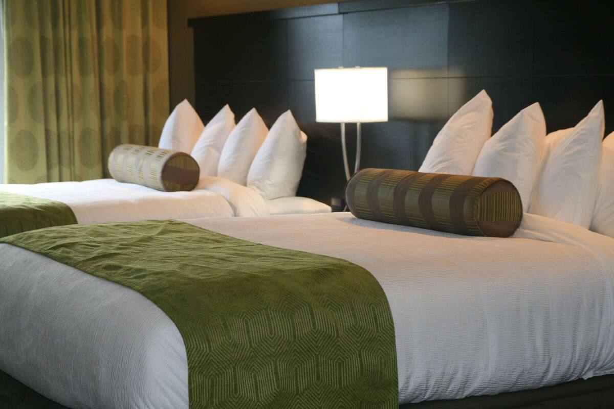 A hotel room with two beds, a large headboard, a bedside lamp, and a window with curtains.