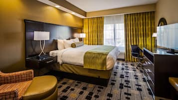 Habitación de hotel con una cama grande, un escritorio con lámpara, una silla, un televisor y una alfombra estampada.