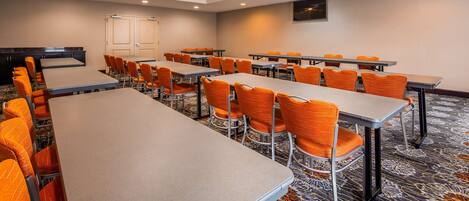A conference room with multiple tables and orange chairs, carpeted floor, and a television on the wall.