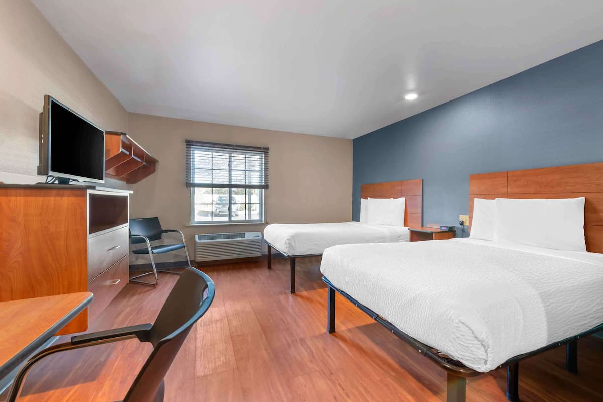 A hotel room with two beds, a desk, a chair, a TV, and a window with blinds.