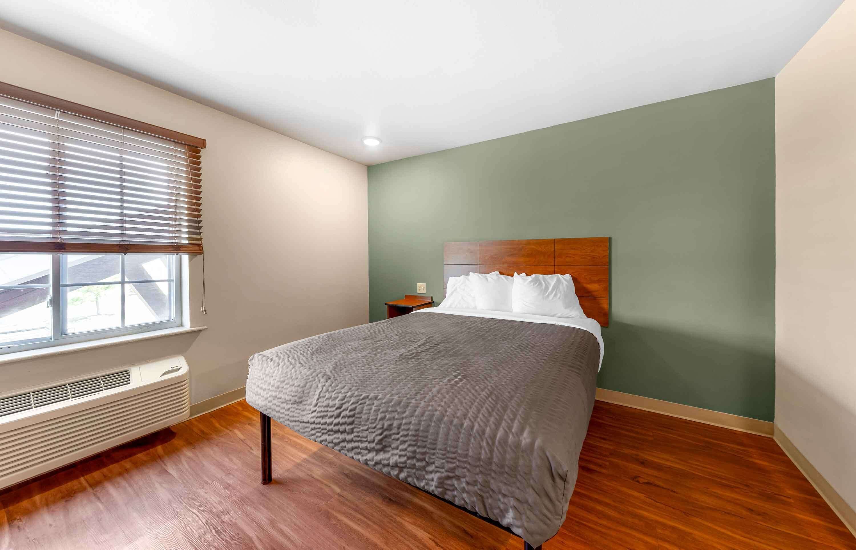 A bedroom with a bed, a window with blinds, a nightstand, and a wall-mounted heater.