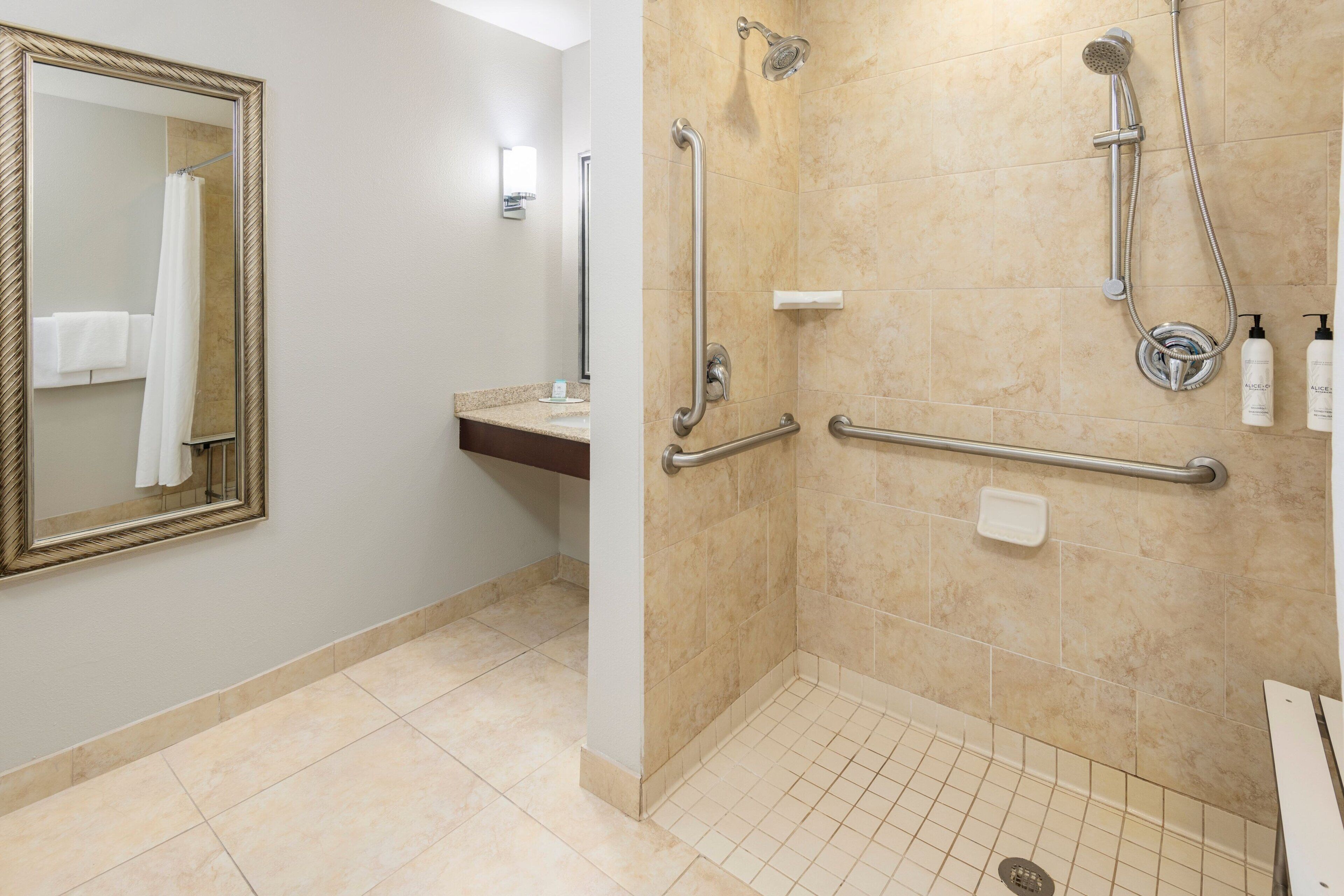 A bathroom with a large mirror, a sink, a shower with grab bars, and a toilet.