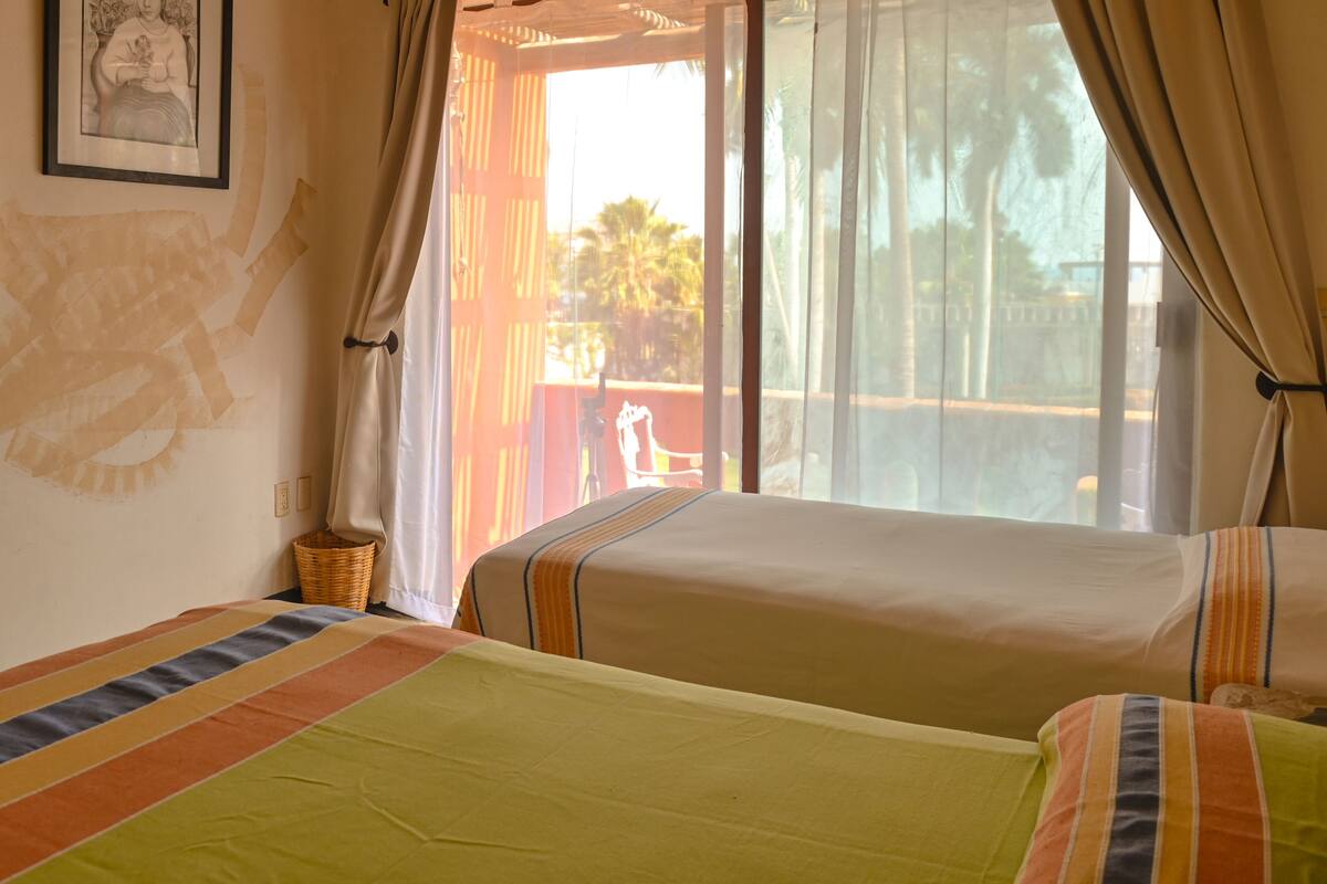 Habitación de hotel con dos camas, una cesta de mimbre y vistas a palmeras tras cortinas translúcidas.