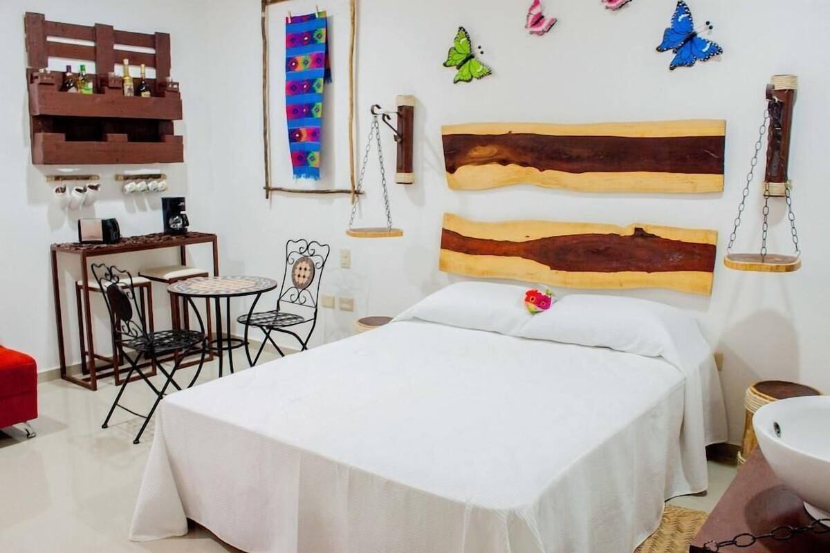 Um quarto com cama, escrivaninha com cadeira, uma mesinha pequena e uma cadeira vermelha. Há itens decorativos nas paredes e uma prateleira de madeira com garrafas.