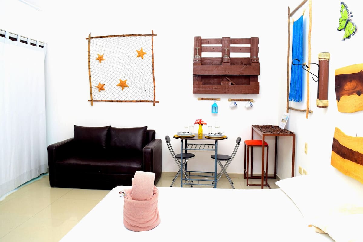 Quarto com sofá marrom escuro, mesa redonda pequena com um vaso e dois copos, cadeira dobrável de metal e toalha rosa sobre uma mesa branca.