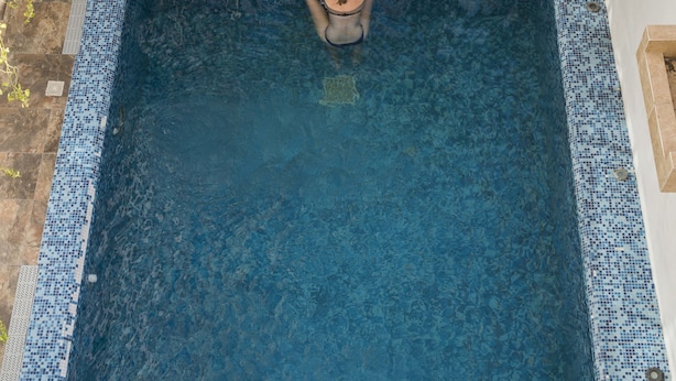 Una piscina en la azotea con dos palmeras, un solárium con baldosas y una zona de estar con mesas y sillas.