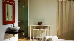 Un baño de hotel con un lavamanos blanco, un espejo con marco rojo, una mesita con dos taburetes, una planta en maceta y una zona de ducha.