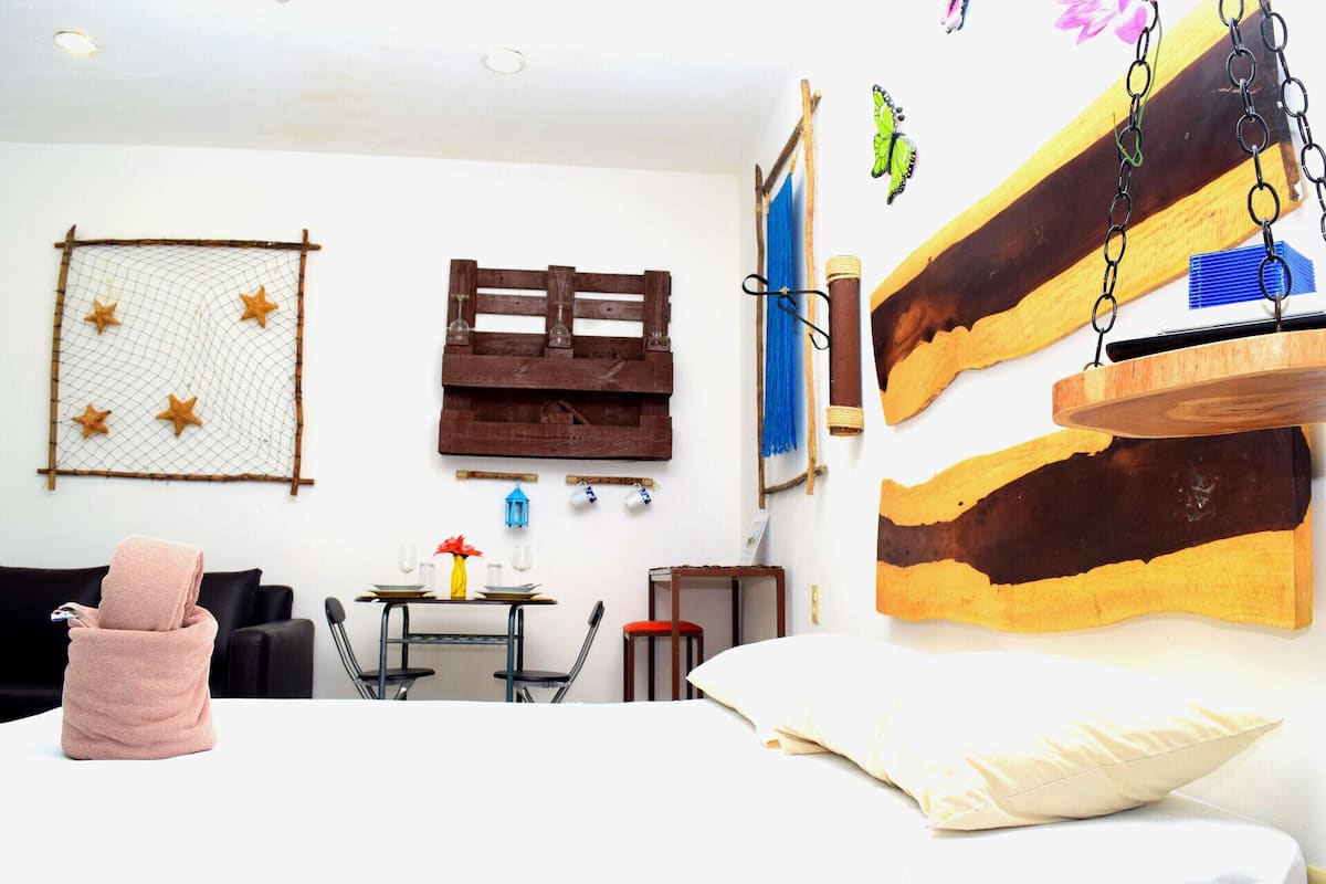 Quarto com cama, uma mesinha e duas cadeiras, luminária pendente e detalhes decorativos na parede, incluindo um palete de madeira e um lanterna azul.