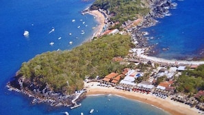 Vista aérea de una zona costera con playa, barcos y un pequeño asentamiento con edificios y un muelle.