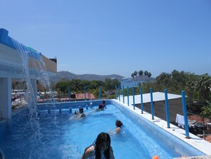 A swimming pool with clear blue water, a water slide, and a view of the ocean and mountains in the background.