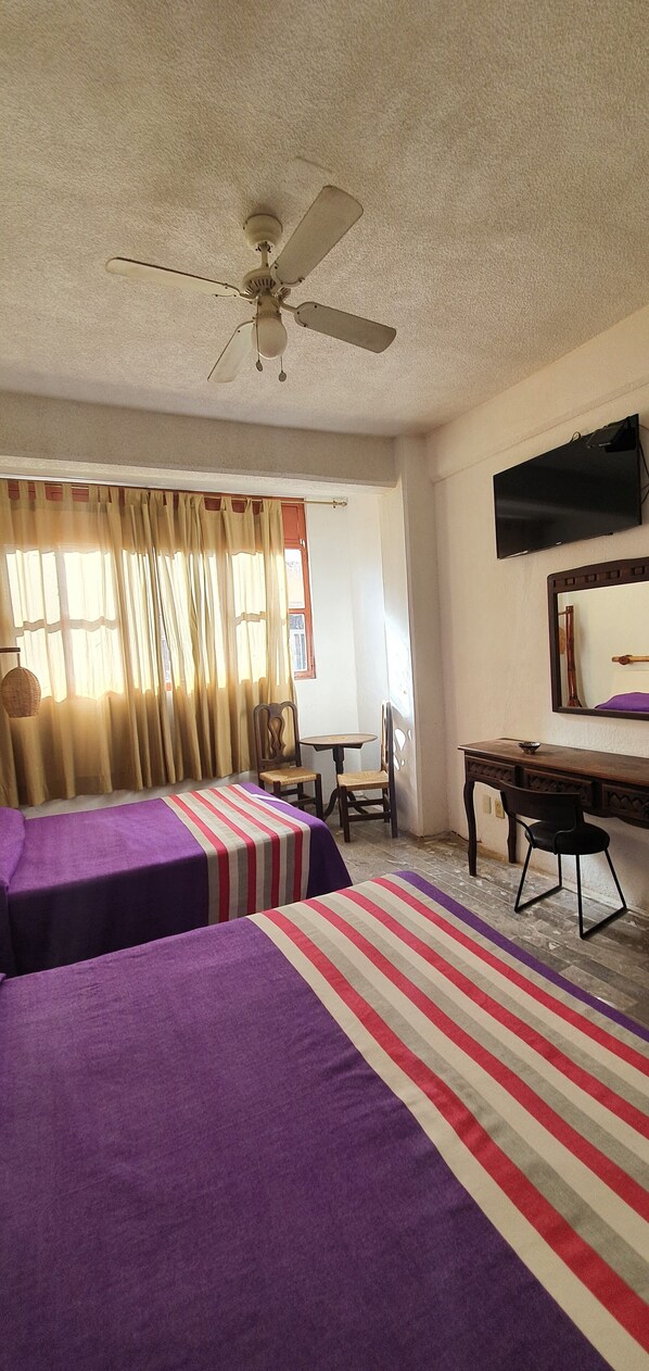 A hotel room with two beds, a desk, a chair, a television, and a ceiling fan.