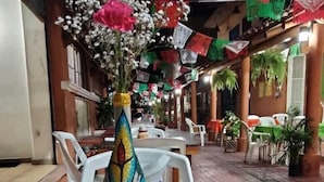 Un área de comedor al aire libre con mesas y sillas, un jarrón decorativo con flores, y banderas coloridas.