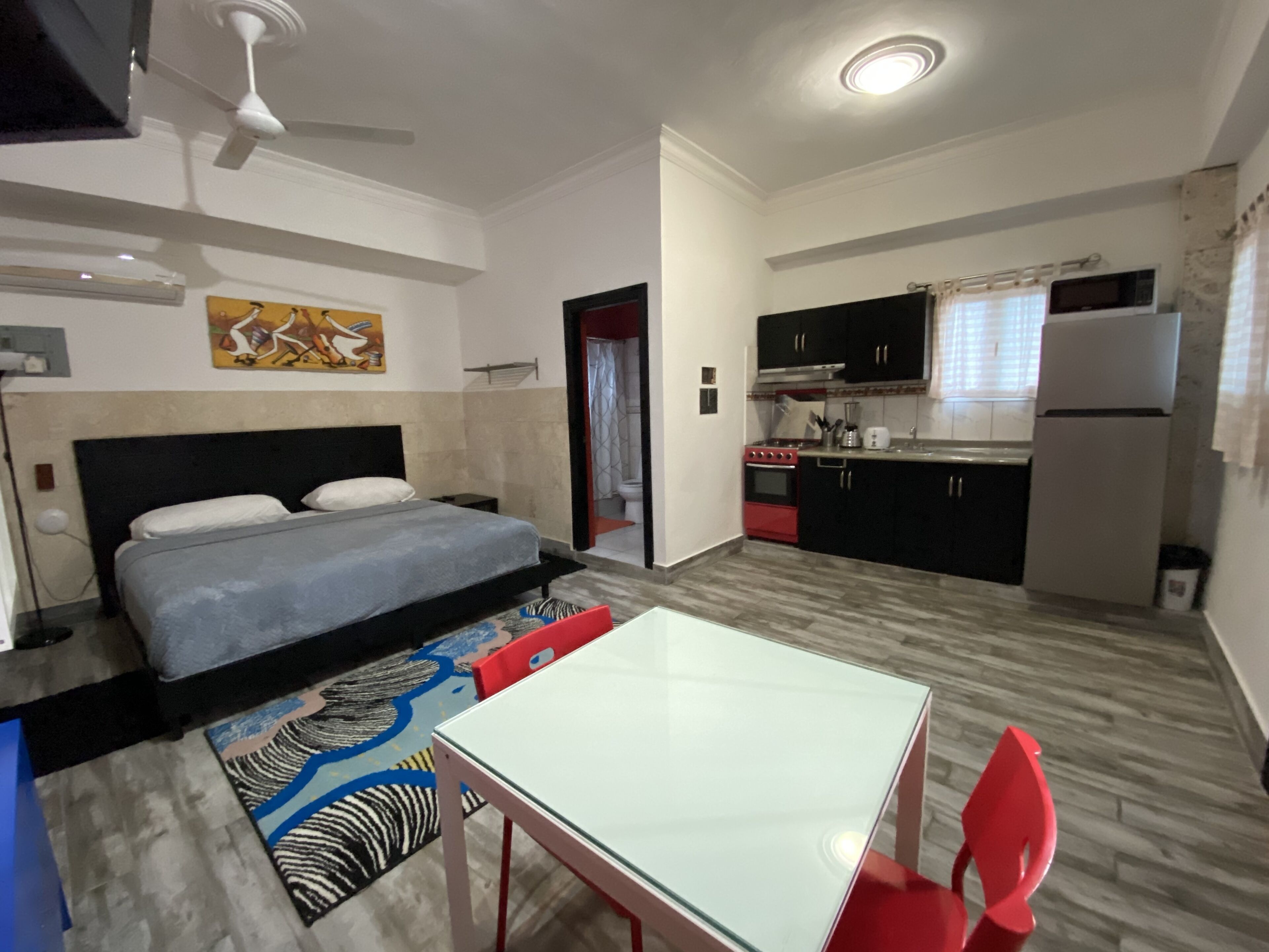 Une chambre à coucher comprenant un lit, une petite table et une chaise. À côté de la chambre se trouve une cuisine équipée d’une cuisinière, d’un four à micro-ondes et d’un réfrigérateur.