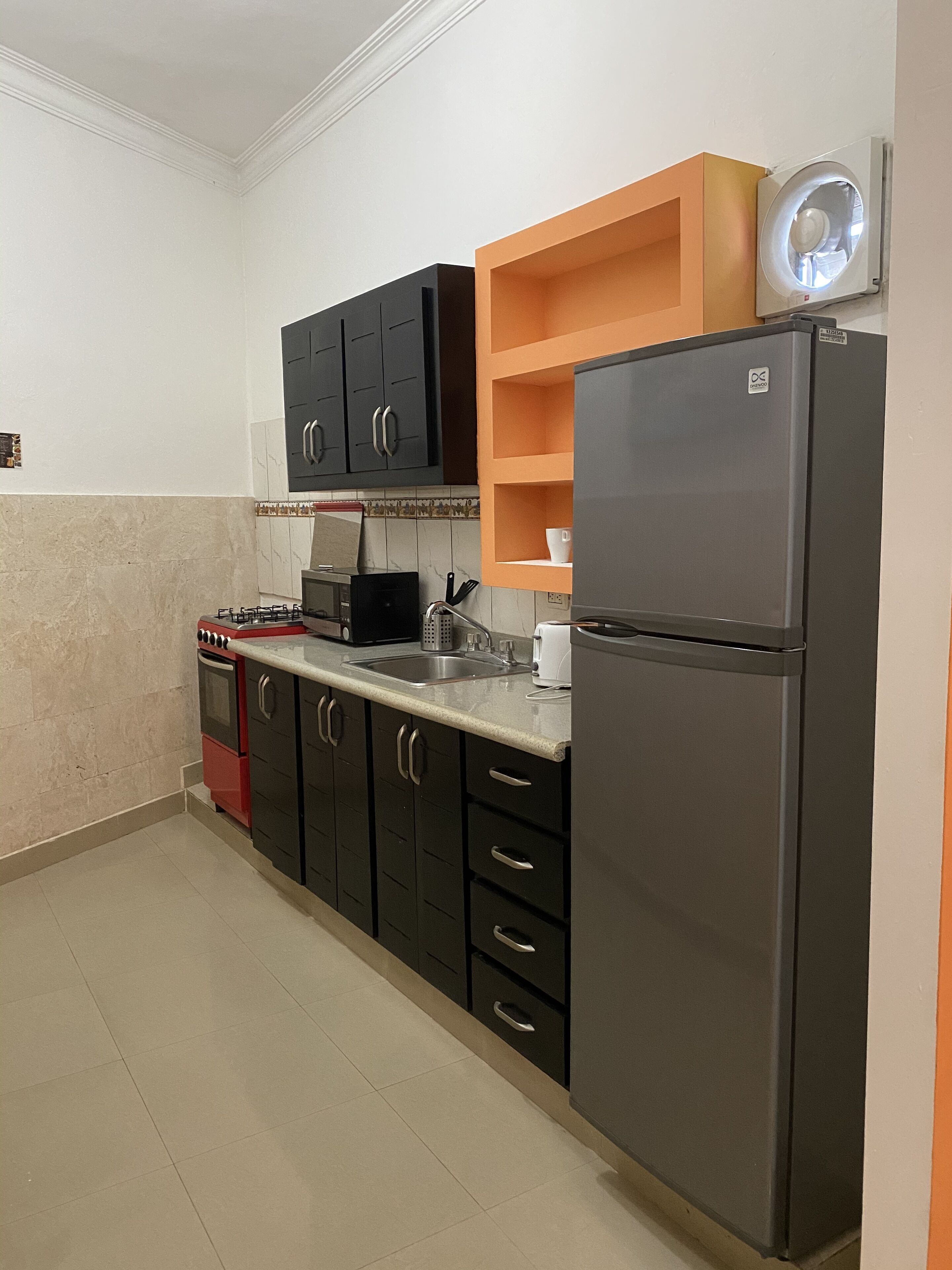Eine Küche mit Gasherd, Backofen, schwarzen Schränken, einem grauen Kühlschrank und orangenen Regalen.