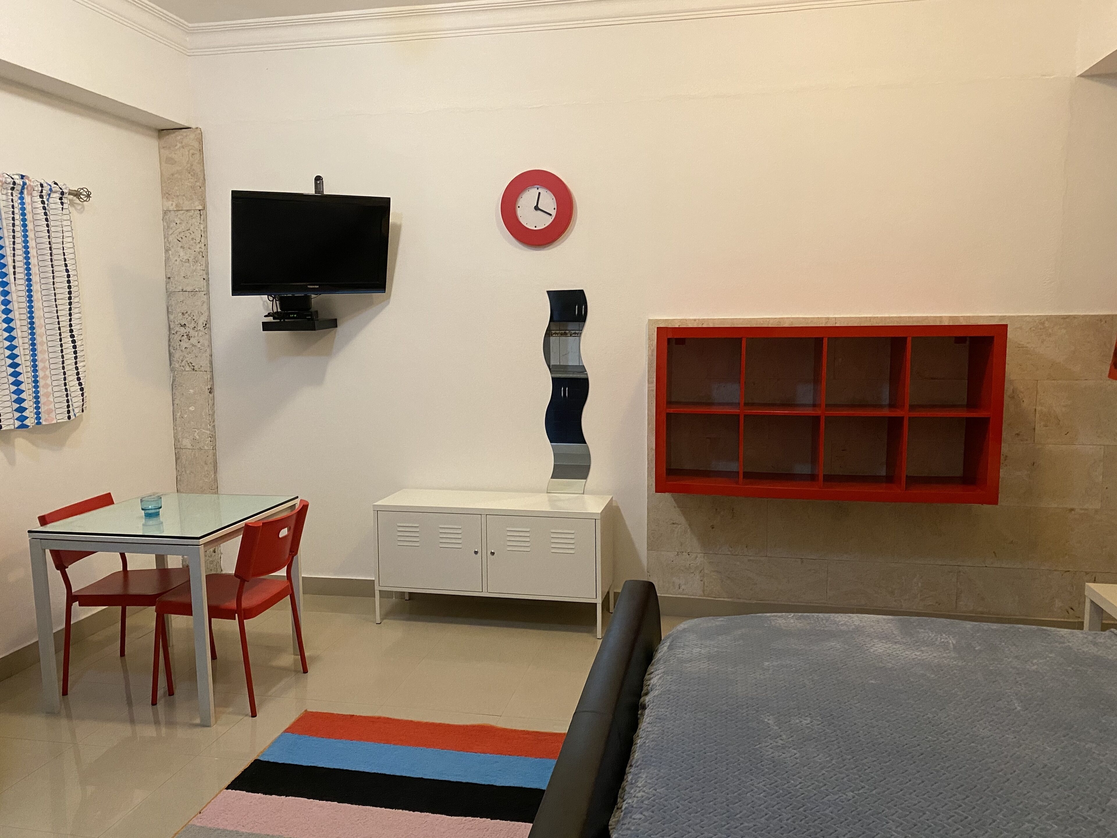 A room with a bed, a TV, a desk with chairs, a red-framed window, and a clock on the wall.