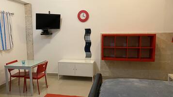 A room with a bed, a TV, a desk with chairs, a red-framed window, and a clock on the wall.