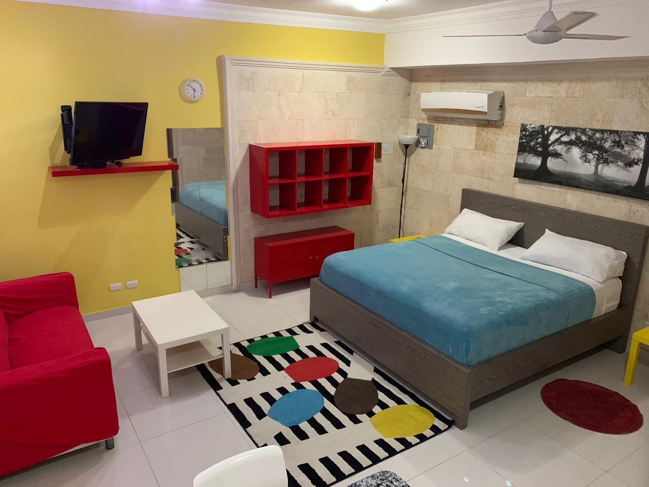 A bedroom with a bed, a red chair, a small table, a TV, a red shelf, and a mirror.