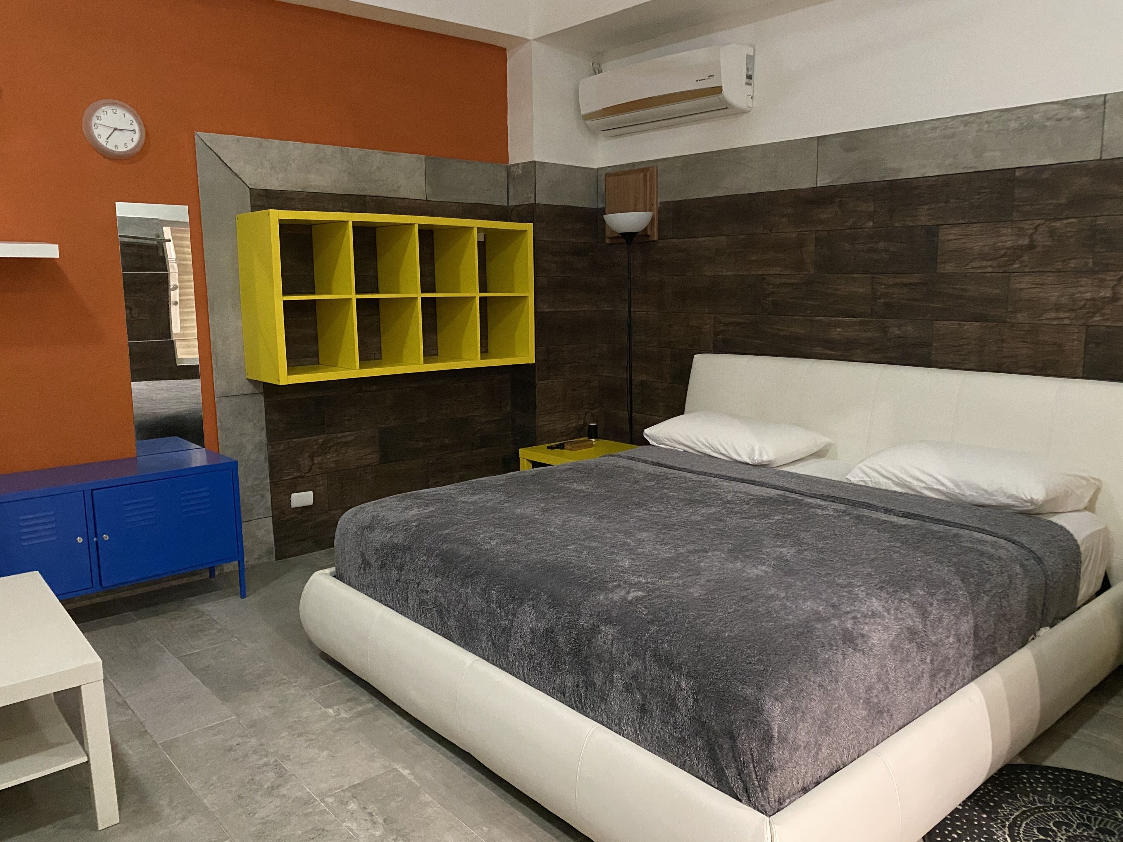 Une chambre moderne avec un lit gris, des oreillers blancs, une étagère jaune, un bureau bleu et une horloge au mur.