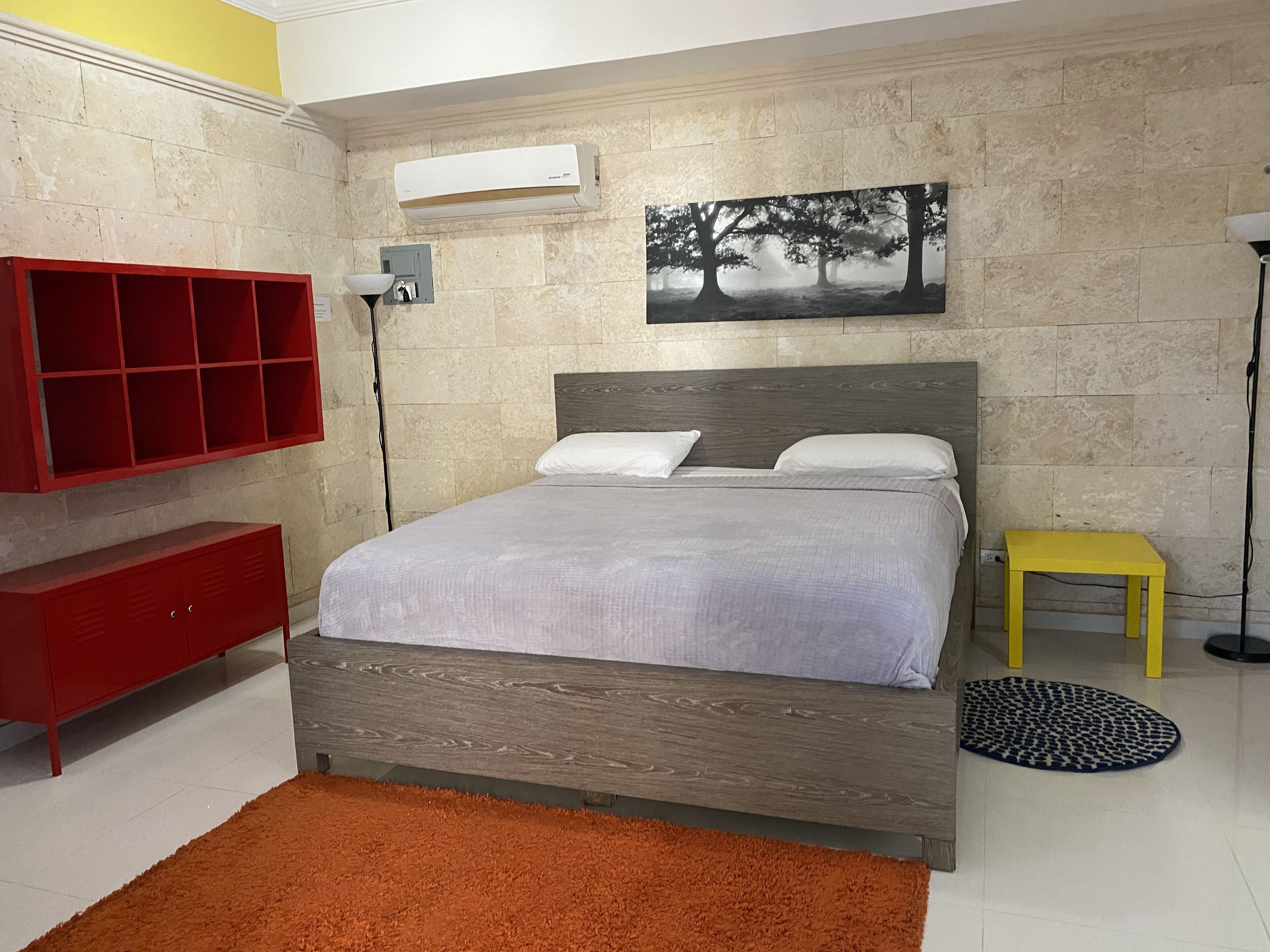 Ein Schlafzimmer mit einem Bett, einem roten Schrank, einem gelben Hocker, einem runden Teppich und einem Bild an der Wand.