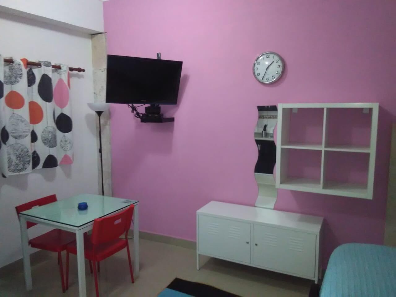 Una habitació amb una paret rosa, un escriptori blanc i una cadira vermella, un televisió fixat a la pared, un rellotge i un armari blanc.