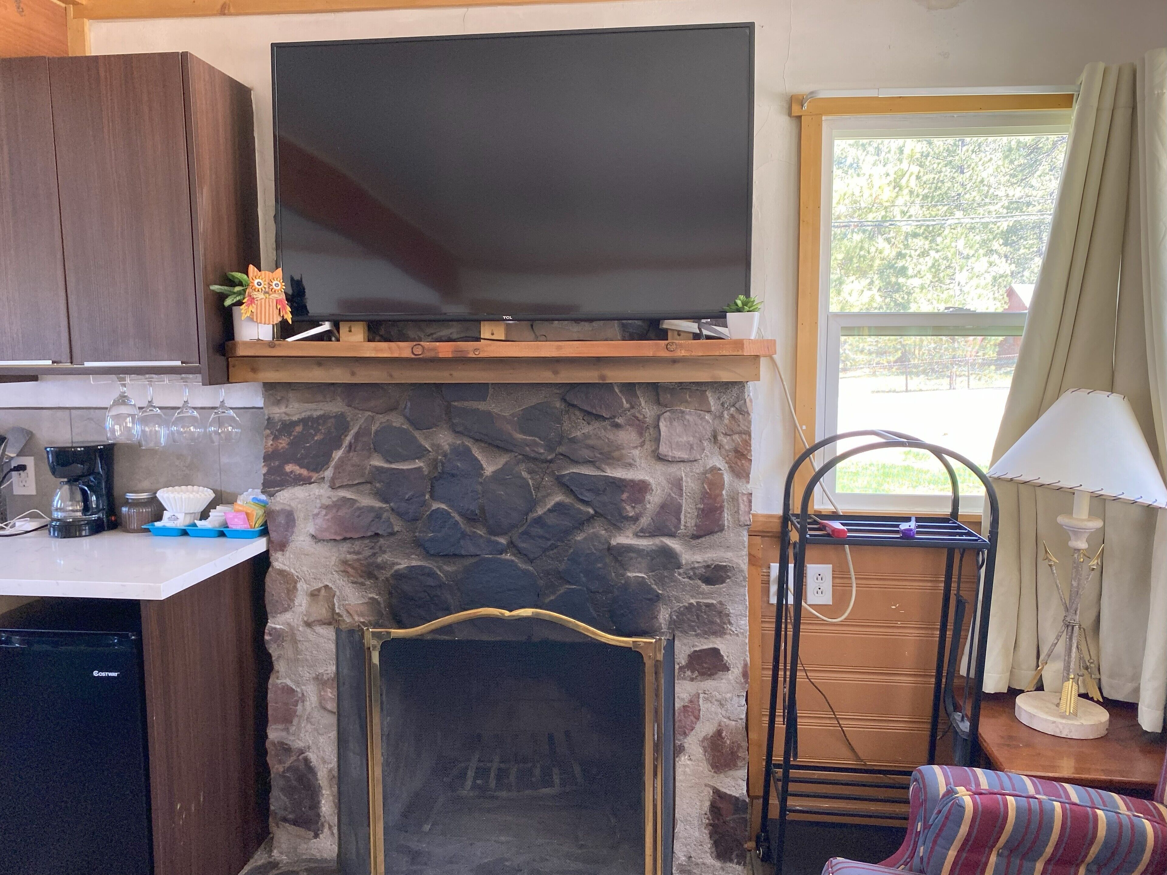 A kitchen with a stone fireplace, a television above it, and a window with curtains.