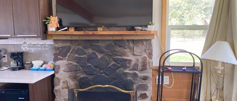 A kitchen with a stone fireplace, a television above it, and a window with curtains.