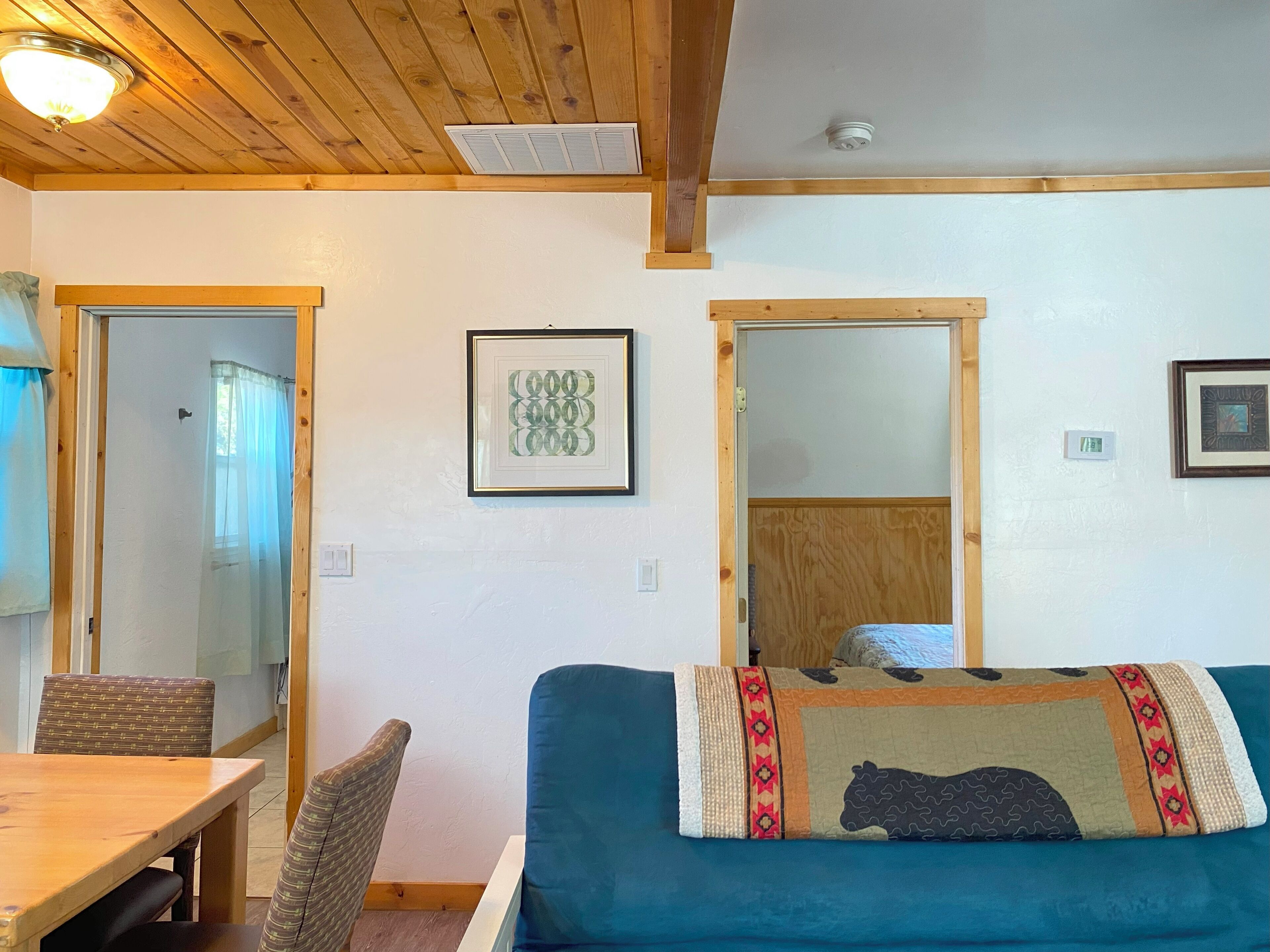 Una stanza con soffitto in legno, una porta con stipite in legno, un'opera d'arte incorniciata, un divano blu con una coperta decorata e un tavolo da pranzo in legno con sedie.