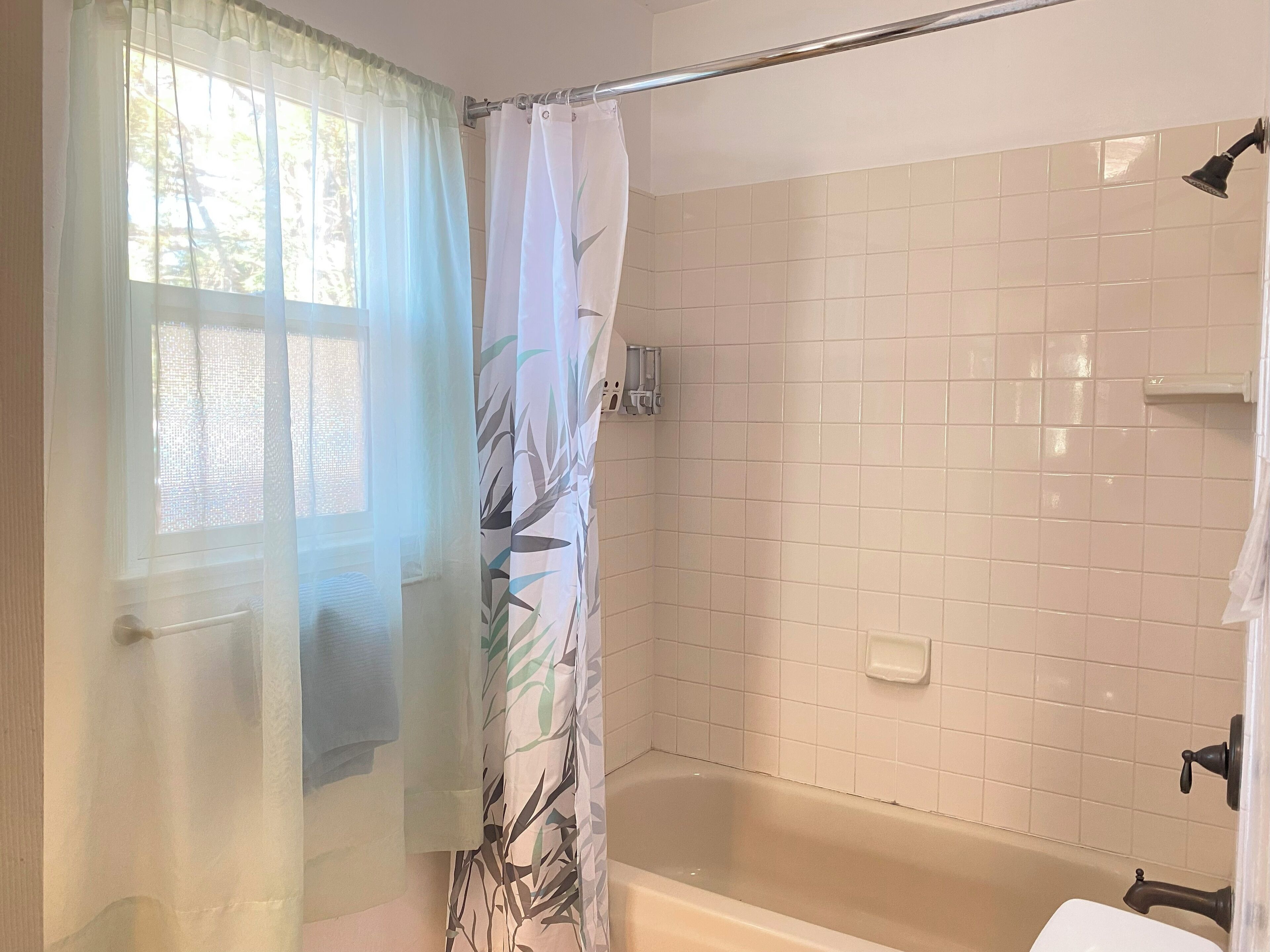 Ein Badezimmer mit Badewanne, Duschbereich und einem Fenster mit durchsichtigen Vorhängen.
