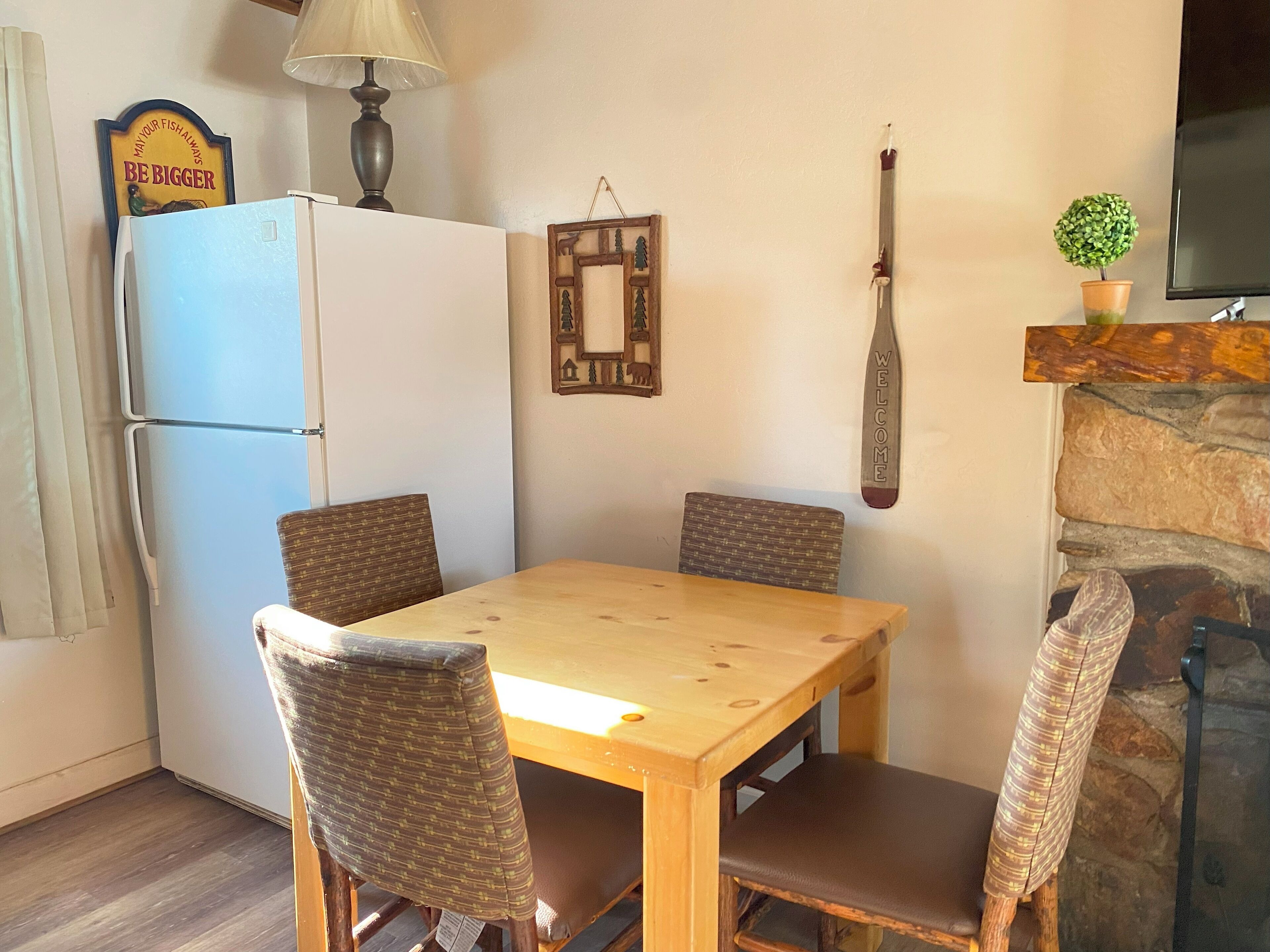 A dining area with a wooden table and four chairs, a white refrigerator, a lamp, a welcome sign, and a small potted plant.