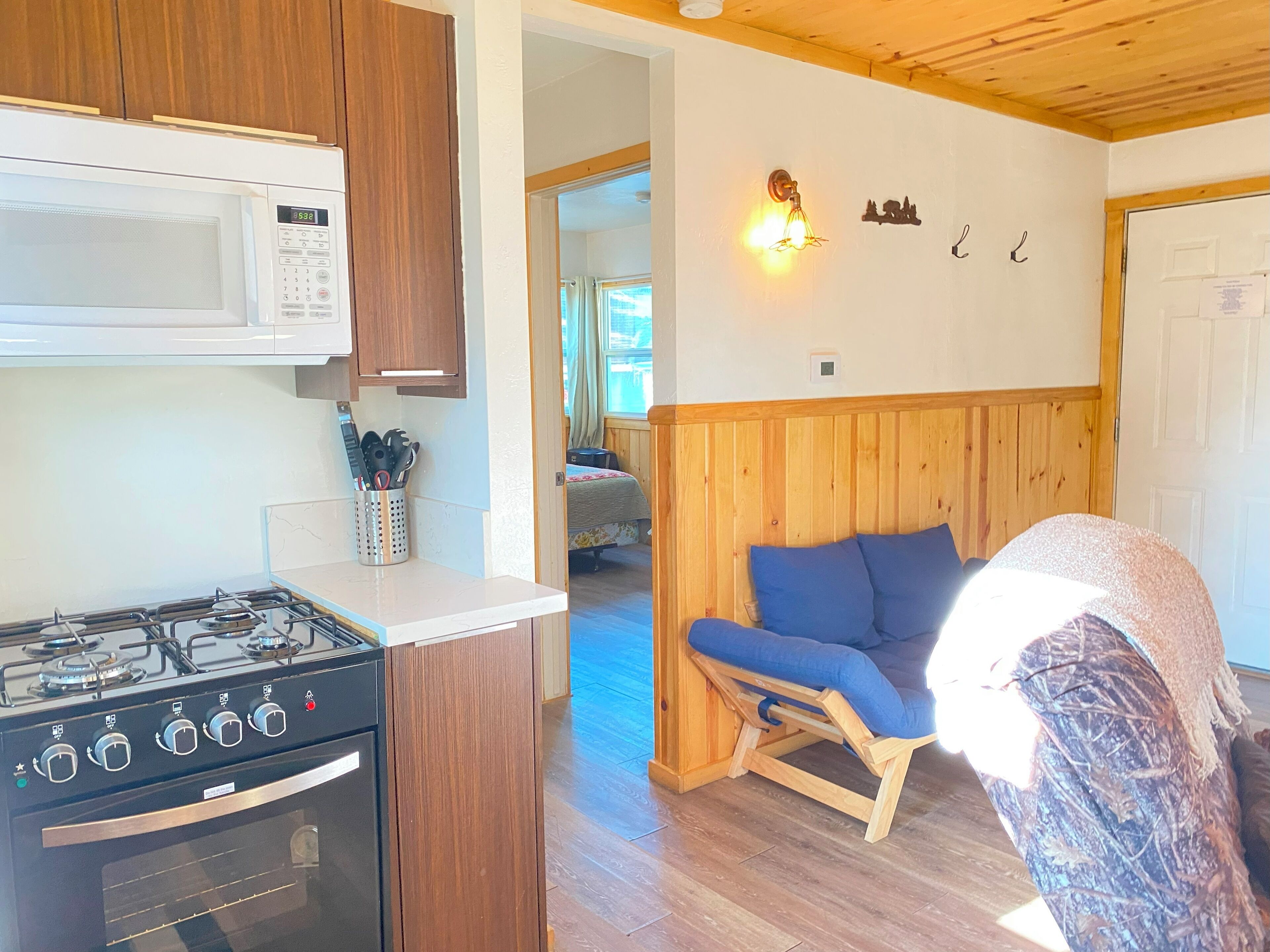 A compact kitchen with a gas stove, microwave, and wooden cabinets. A small seating area with a blue cushion and wooden frame is visible.