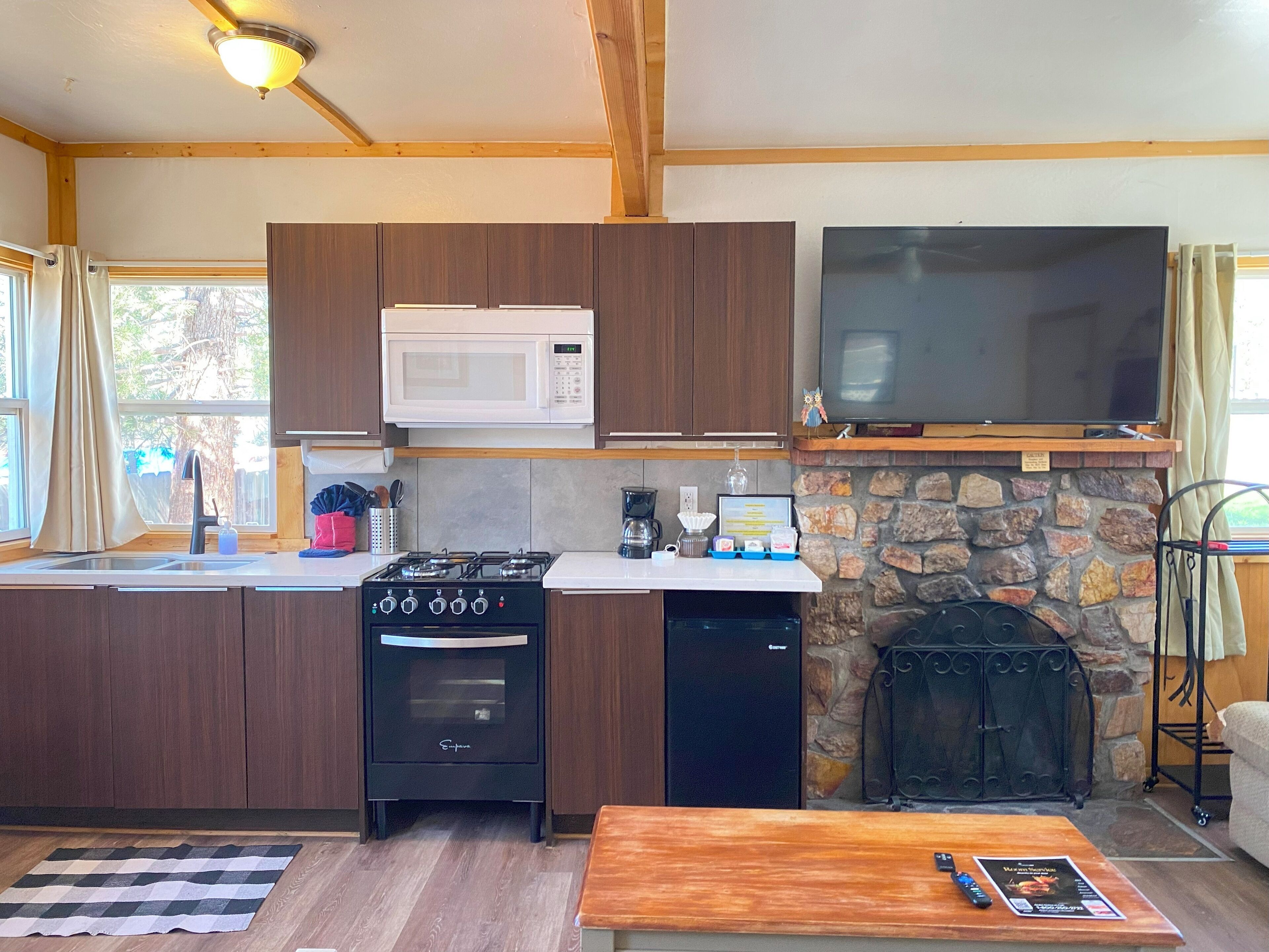 A compact kitchen with a stove, microwave, and oven, a stone fireplace, a flat-screen TV, and a dining area with a table.