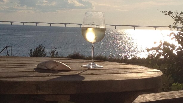 A glass of white wine on a wooden table with a view of a bridge over water and the sun setting in the background.