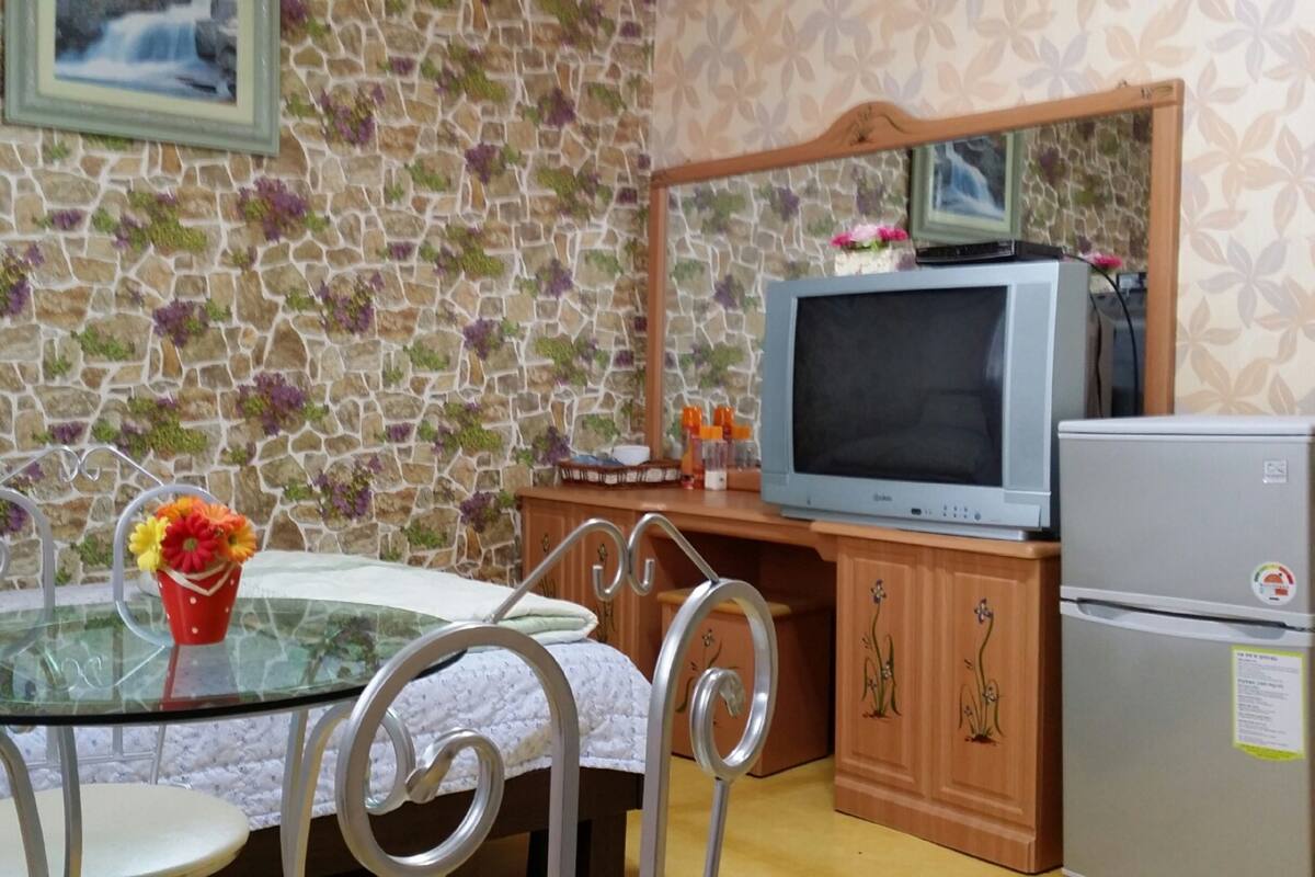 Habitació petita amb paret de pedra, taula de vidre i cadires metàl·liques, televisió i nevera.