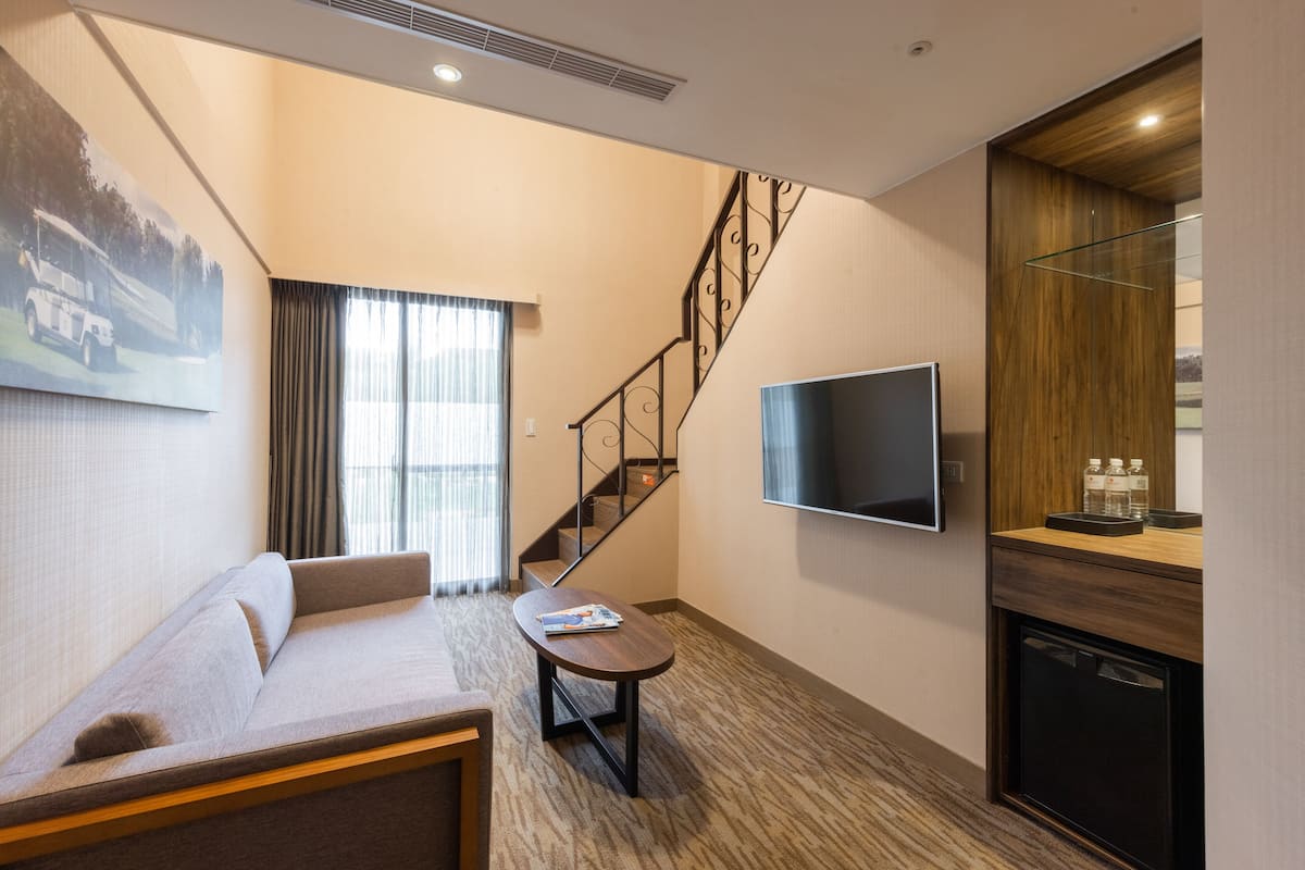 A modern hotel room with a sofa, a small table, a flat-screen TV, a wooden cabinet, and a staircase.