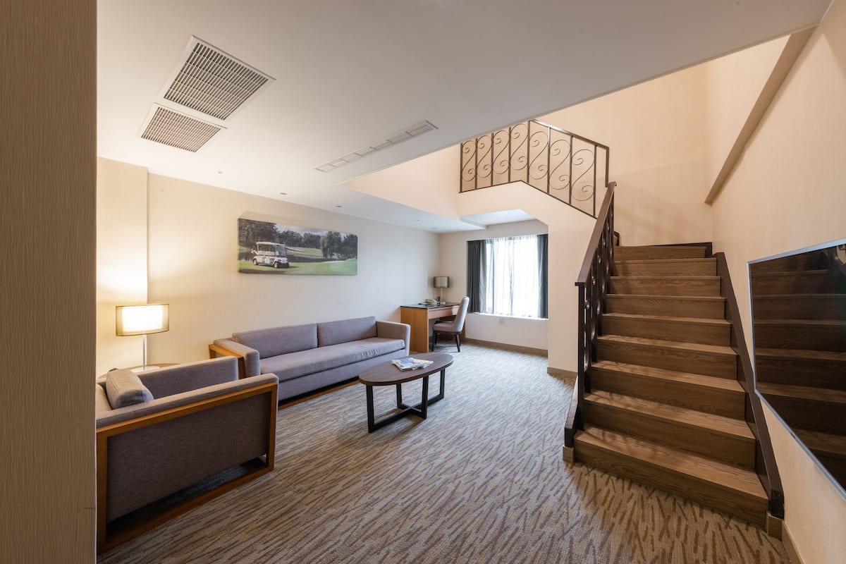 A hotel room with a sofa, armchair, coffee table, desk, and staircase.