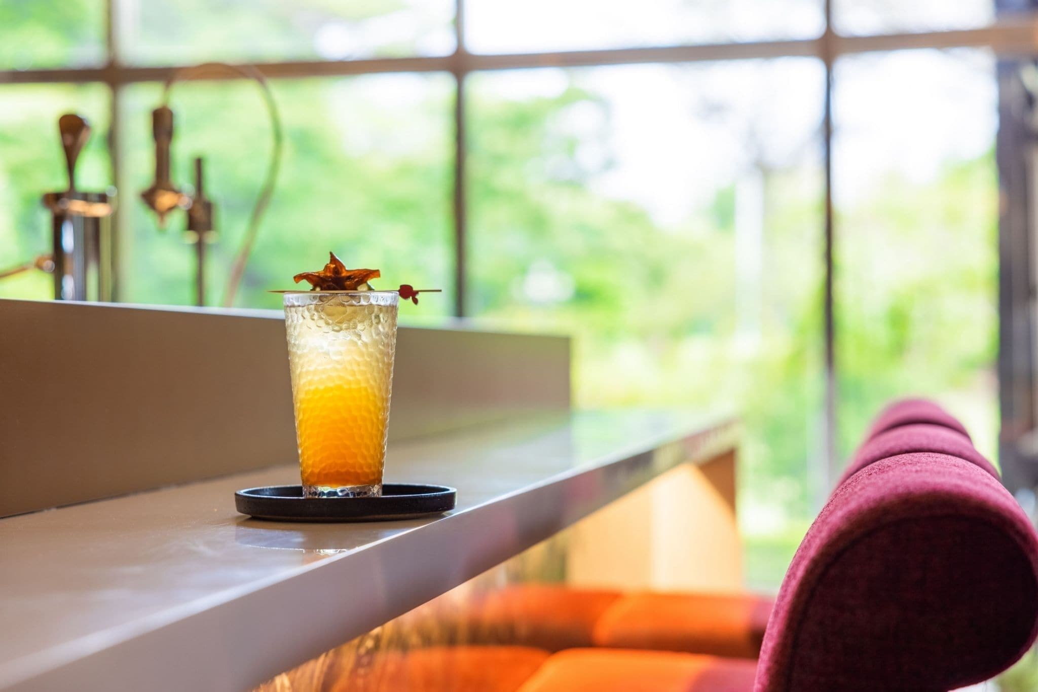 A glass of iced beverage with a garnish on a coaster, placed on a window sill with a view of greenery outside.
