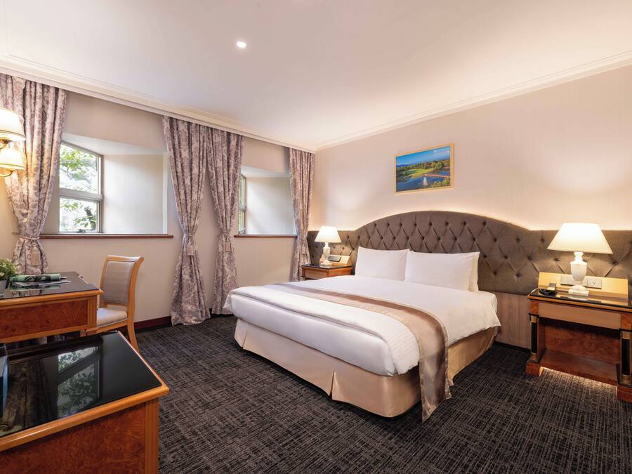 A hotel room with a large bed, two bedside tables with lamps, a desk with a chair, and a window with curtains.